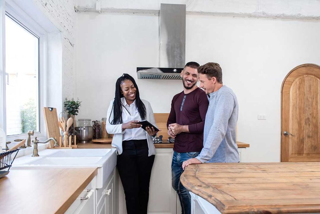 A couple reviews a real estate agreement with an agent at a kitchen table beside a notebook and house documents.