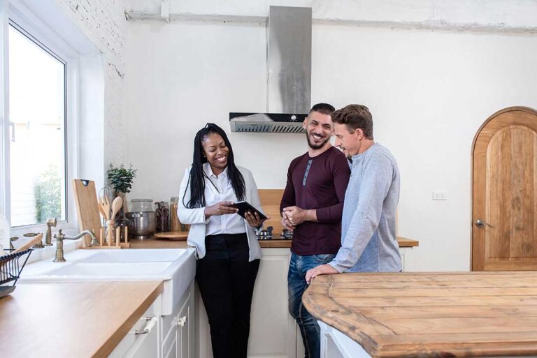 A couple reviews a real estate agreement with an agent at a kitchen table beside a notebook and house documents.